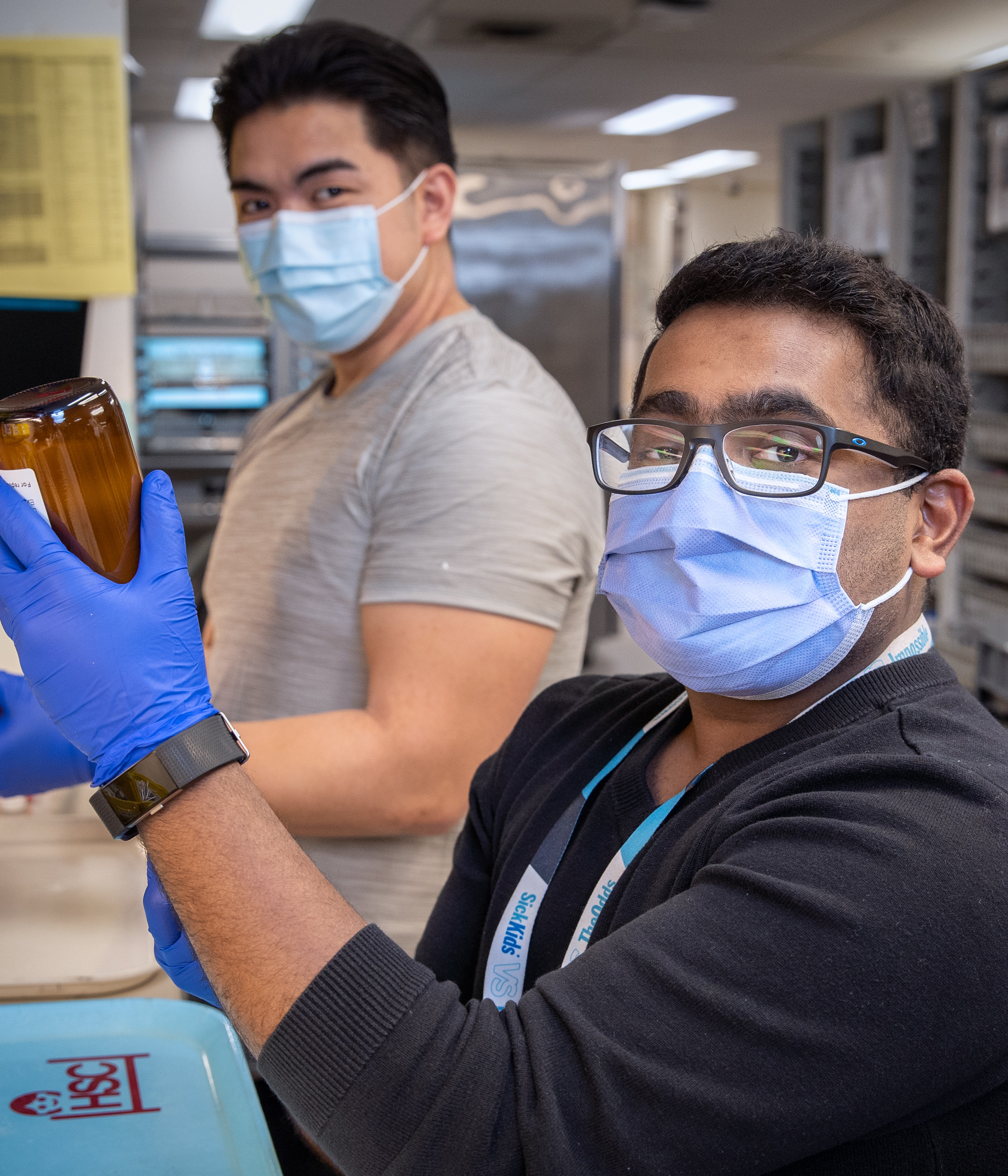 Two members of the SickKids pharmacy team are shown in a lab, with one individual holding up an item to the camera.