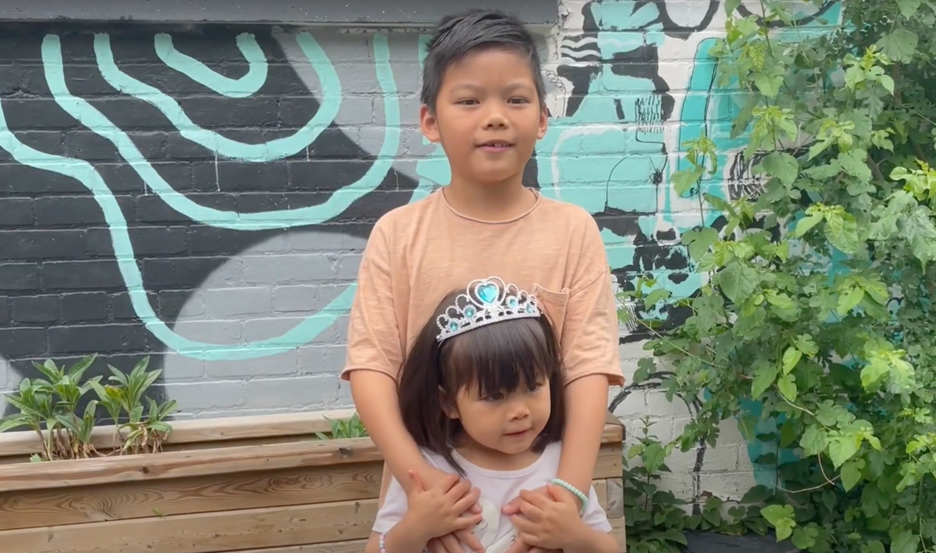 In this preview image of a video, a little girl with a crown is being hugged from behind by her brother. Both kids are outdoors in front of a wall with a mural and some green leaves on a bush or tree.