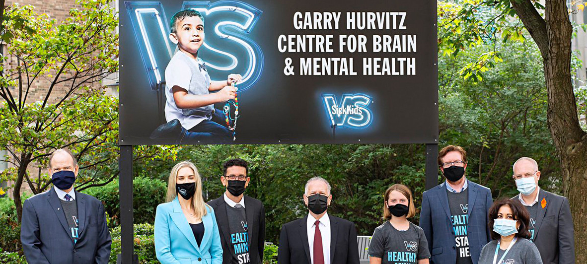 Members of SickKids as well as Garry Hurvitz stand in formal business attire in front of a sign that reads, Garry Hurvitz Centre for Brain & Mental Health. Everyone is wearing a face mask and standing outdoors in front of greenery.