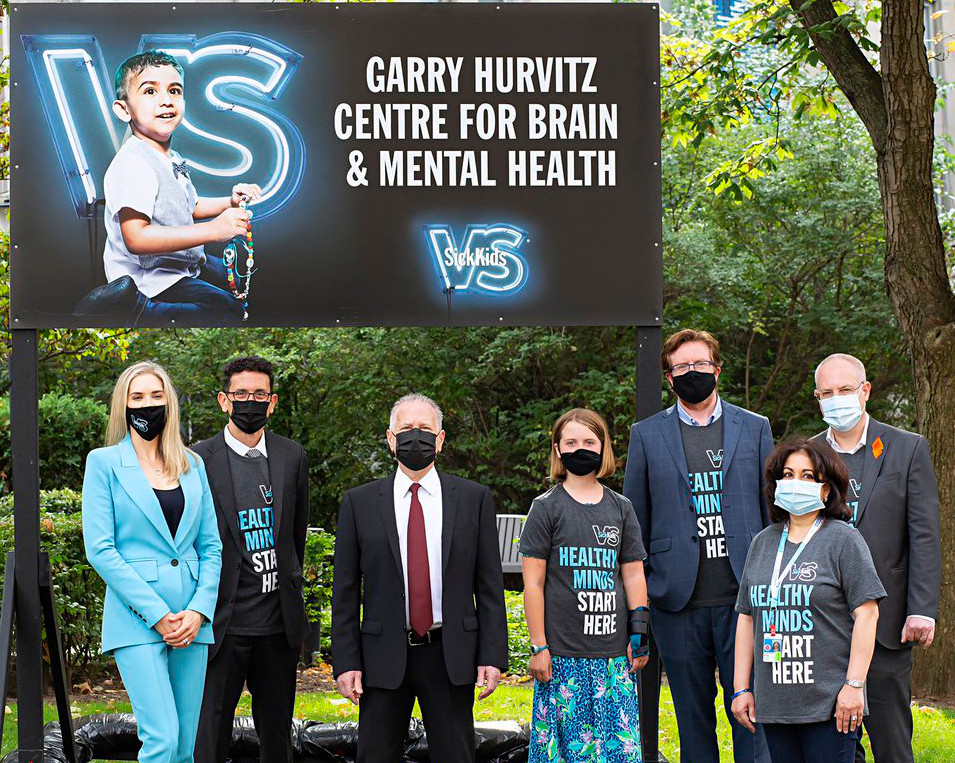 Members of SickKids as well as Garry Hurvitz stand in formal business attire in front of a sign that reads, Garry Hurvitz Centre for Brain & Mental Health. Everyone is wearing a face mask and standing outdoors in front of greenery.