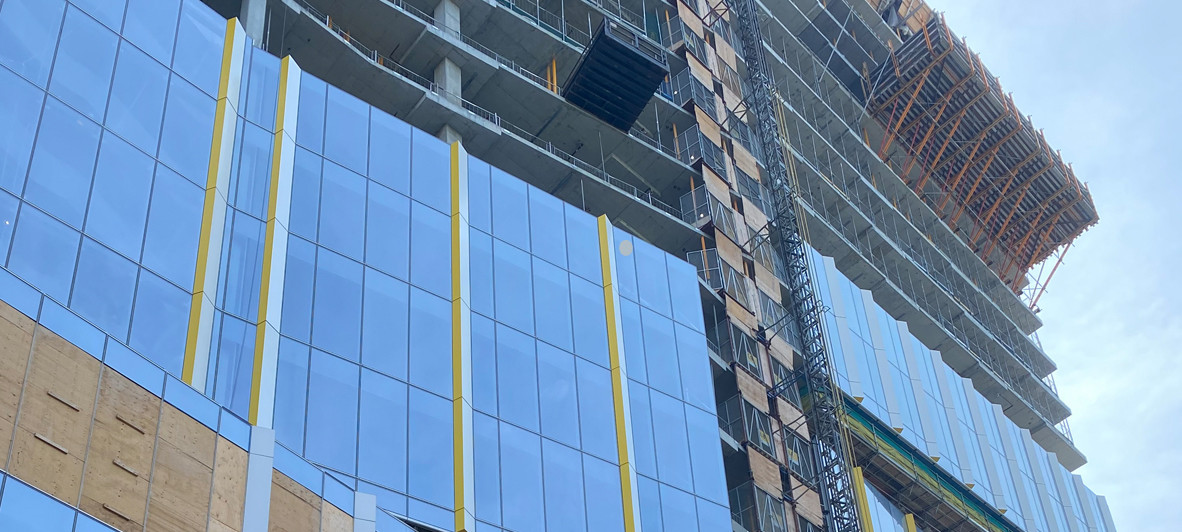 The Patient Support Centre is shown under construction with blue glass and yellow trim partially installed on the bottom portion of the building, and still in the early stages of construction toward the top.