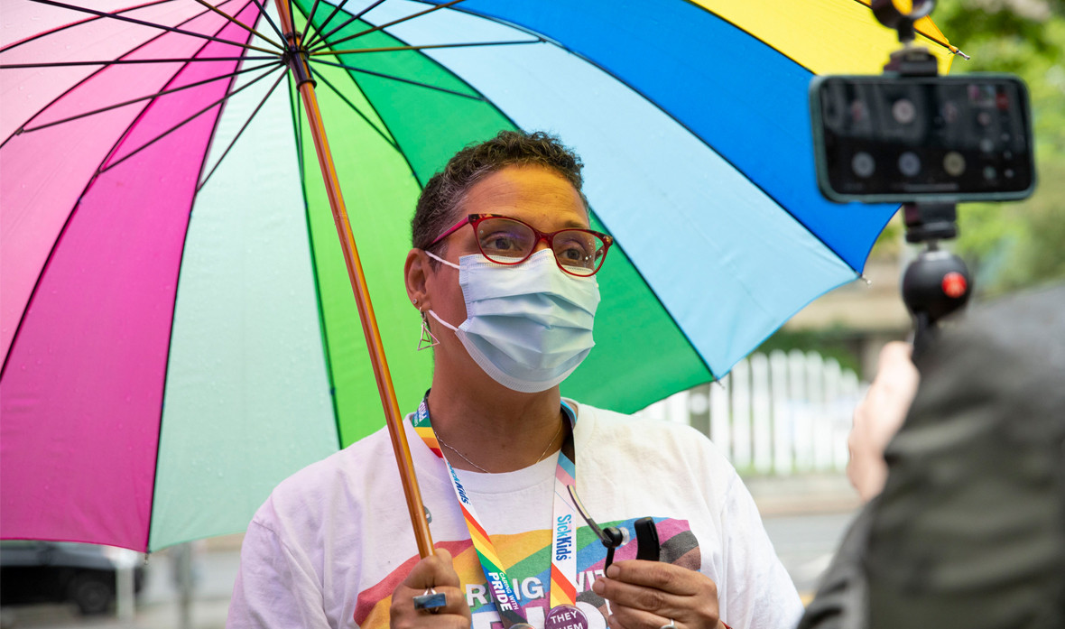 Tee Garnett stands underneath a rainbow-coloured umbrella with a face mask while getting their photo taken at a SickKids Pride event.