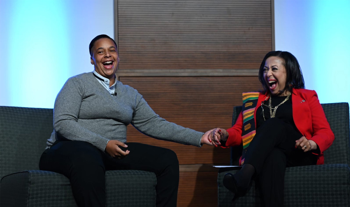 Raia Carey from SickKids Foundation is shown seated in a armchair while laughing and reaching out to hold the hand of her mother, Rosemary Sadlier, who is also laughing and holding Raia's hand.