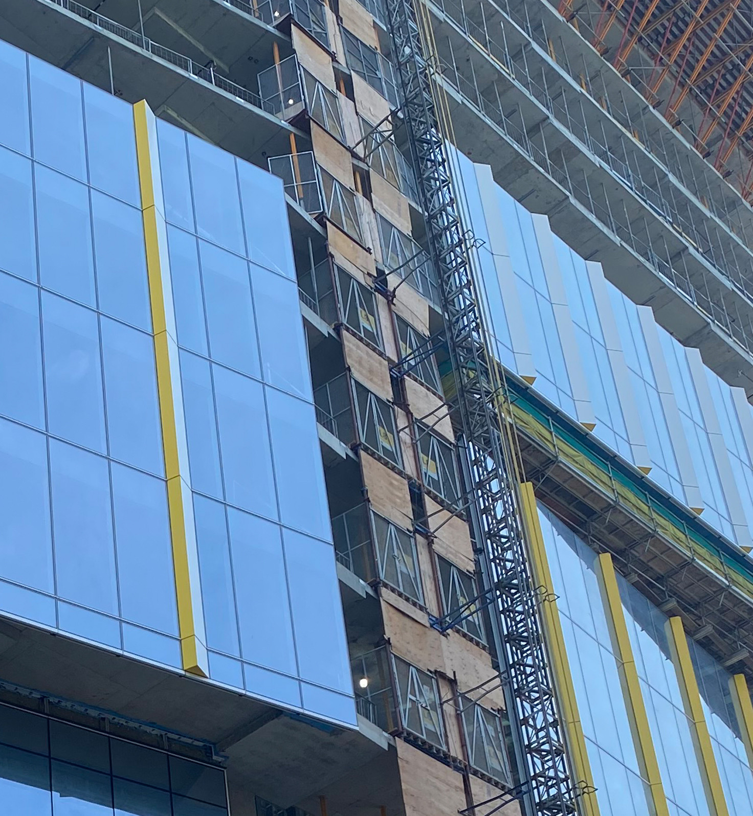 The Patient Support Centre is shown under construction with blue glass and yellow trim partially installed on the bottom portion of the building, and still in the early stages of construction toward the top.