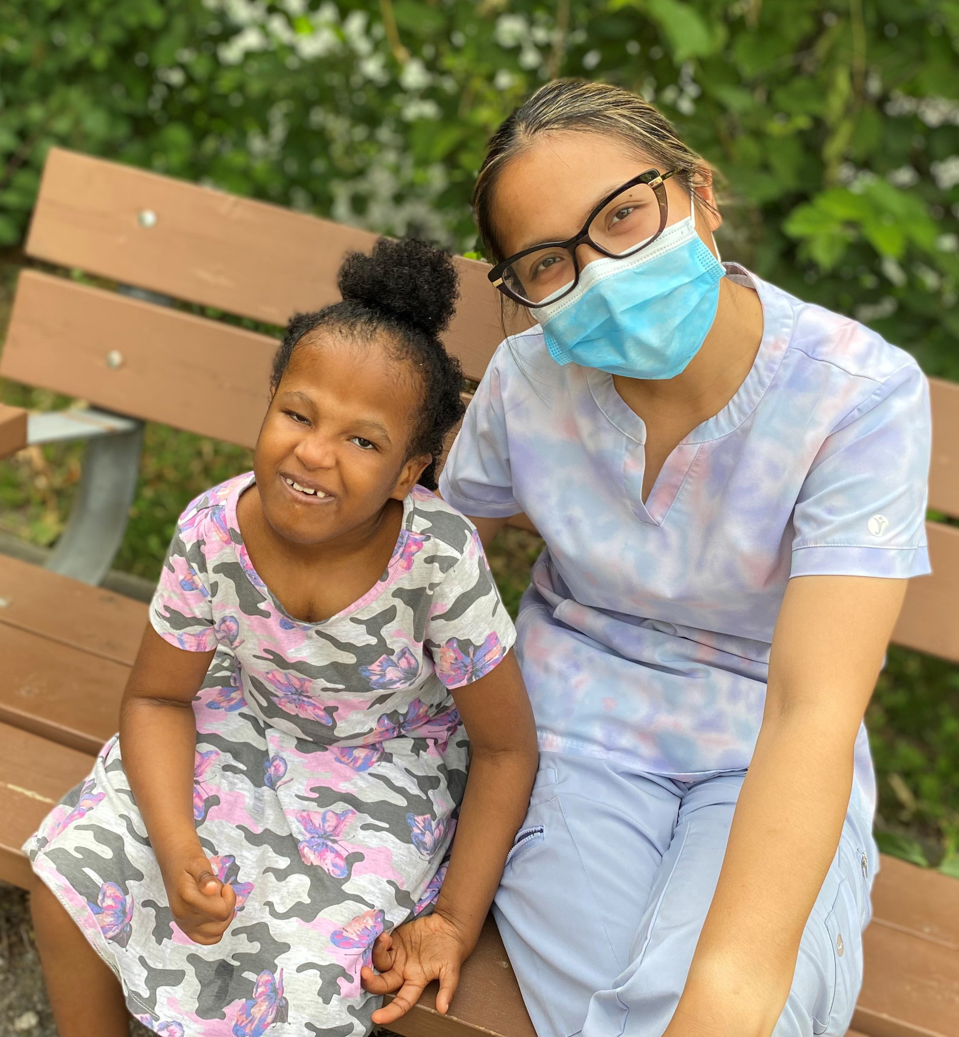 A smiling young child appears to the left of an adult who is wearing a face mask. They are both seated on a wooden bench.