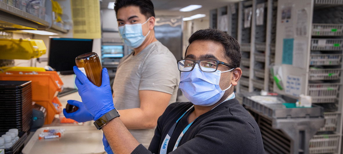 Two members of the SickKids pharmacy team are shown in a lab, with one individual holding up an item to the camera.