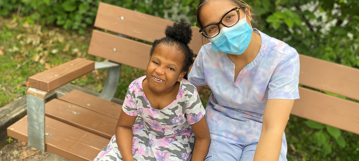 A smiling young child appears to the left of an adult who is wearing a face mask. They are both seated on a wooden bench.