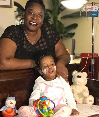 A young child named Jahlisa is seated next to a couple of toys and a stuffed bear, slightly supported by the hand of her smiling mother, Sophia, who is directly behind her.