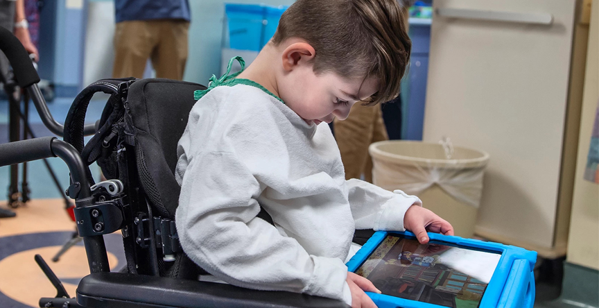Child sits in wheelchair playing with tablet.