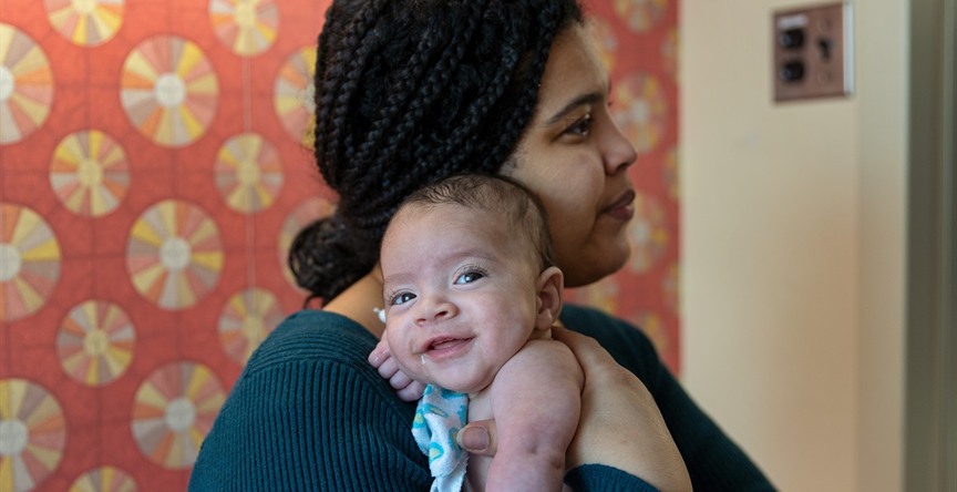An adult holds a small baby against their shoulder in a room with patterned wallpaper