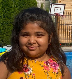 A portait of Ayla. She has brown skin and shoulder length brown hair. She wear a yellow floral dress and is standing infront of a pool with a baseketball net visible in the background.