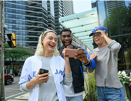 Three people outside in downtown Toronto, wearing Blue Jays merchandise, laughing and looking at a phone screen together