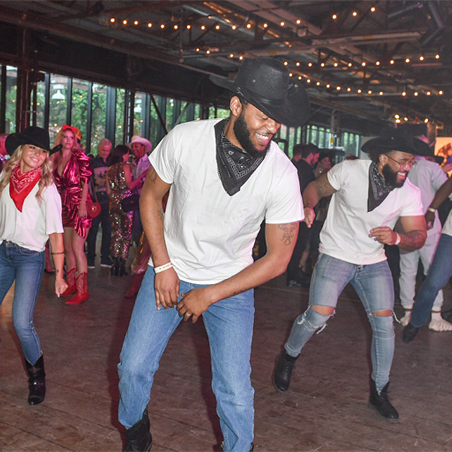 A group of several people smiling while dancing in formation at Scrubs in the City Nashville. They are wearing blue jeans, white T-shirts, cowboy boots, cowboy hats and scarves. 