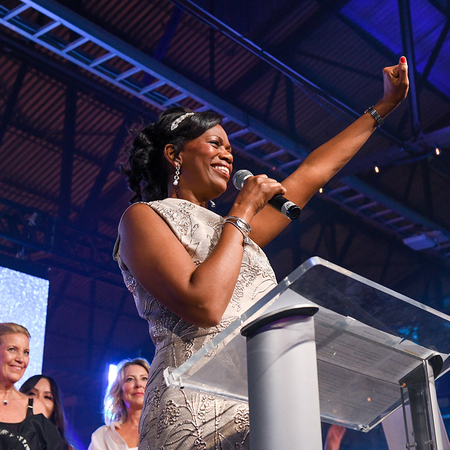 SickKids CEO Jennifer Bernard greets the crowd with a microphone in her right hand and left arm raised with a closed fist. She is wearing a white sleevless dress, long earrings and a headband.