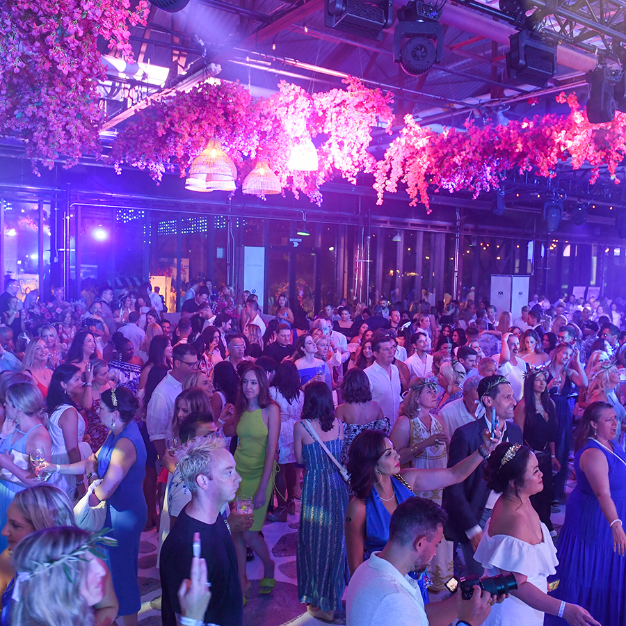 A large crowd of well dressed party goers mingle with eachother. There are pink plants dangling from above.