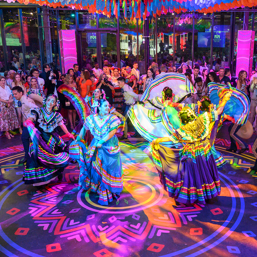 A large crowd of well dressed party goers mingle with eachother watch dancers perform a skirt dance.