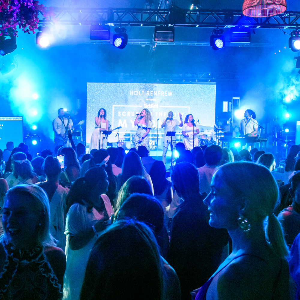  An Image of a band, dressed in white playing for a large crowd of Scrubs 2023 party attendees The band has 4 singers, someone on saxophone and someone playing the keyboard.